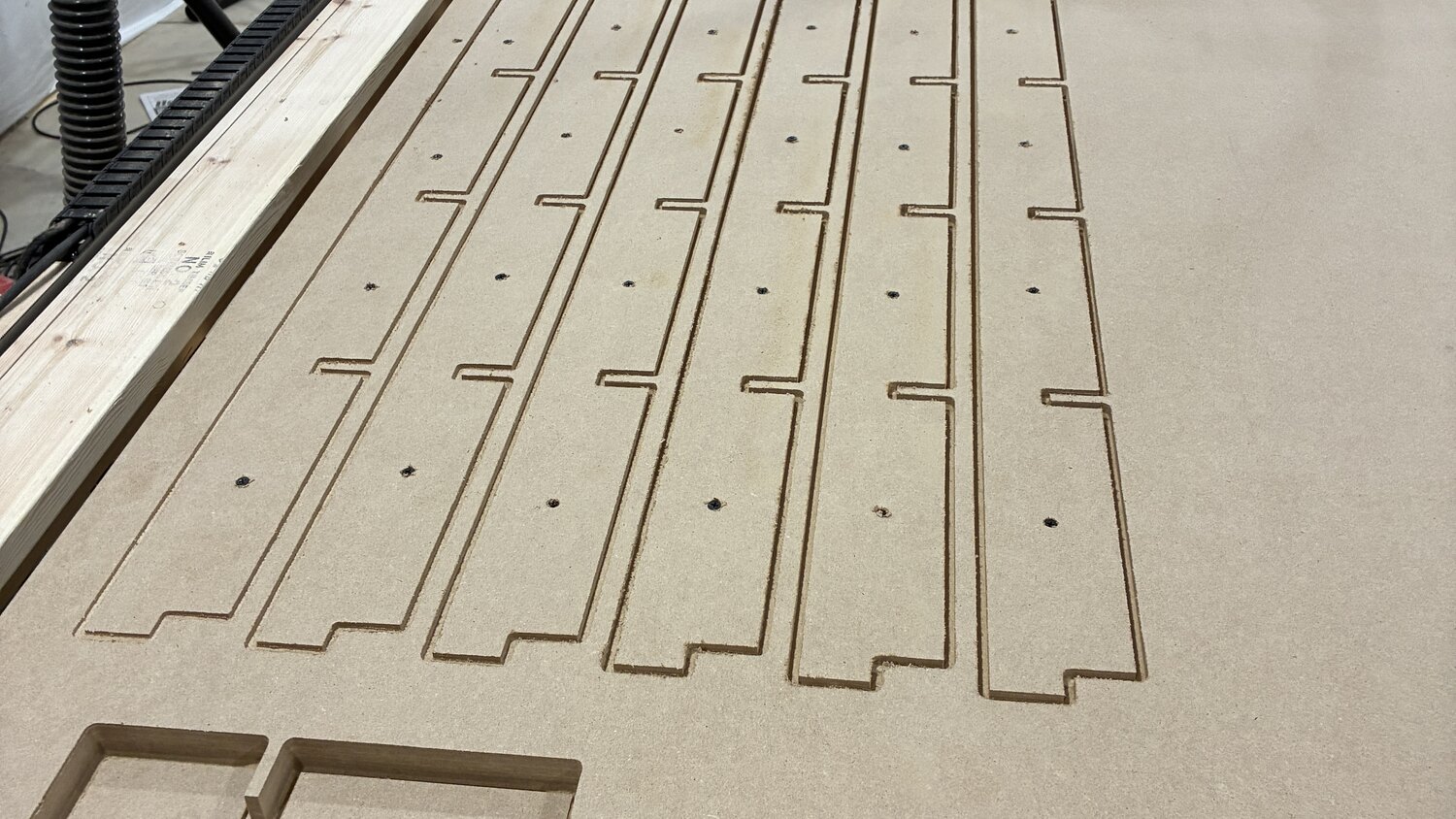 Long ribs cut into a sheet of MDF on the CNC bed, each with a row of slots and stepped ends where they splice together