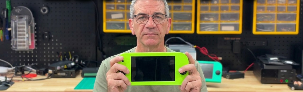 A handheld CNC pendant held in two hands in a workshop. The case is bright green with an analog thumbstick on each side and a seven inch touchscreen in the middle, the same screen used in the Fireside panel. Behind it is a workbench with a pegboard of tools and yellow parts bins.