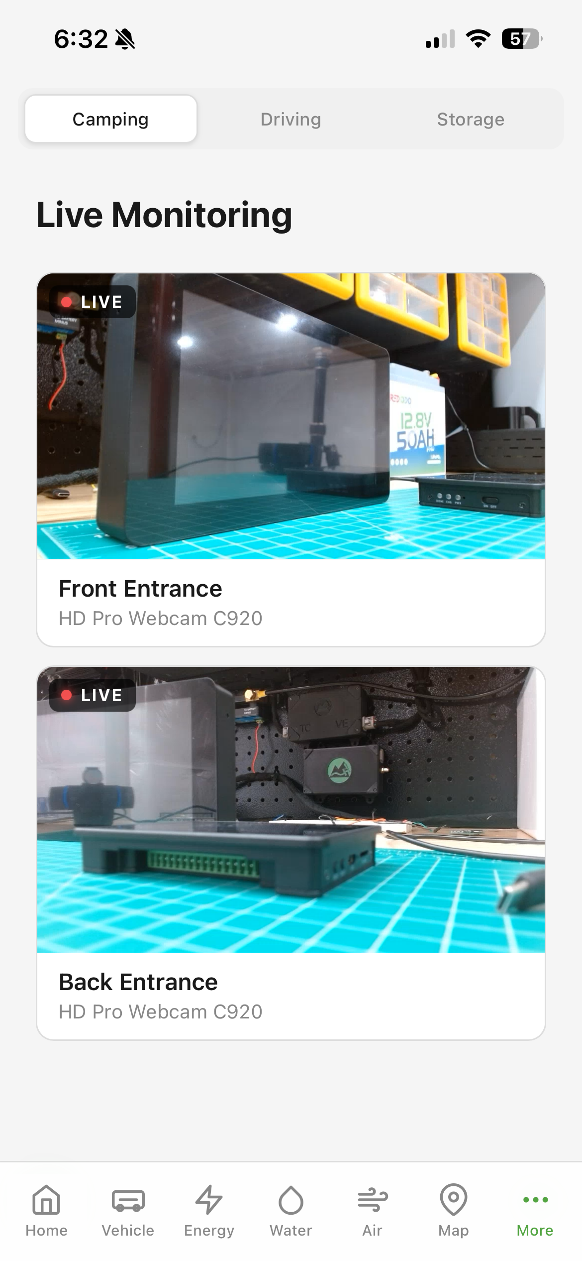 Overlook on an iPhone showing the Live Monitoring page. Two live camera tiles are stacked, each with a red LIVE badge in the corner. The top tile is labeled Front Entrance, HD Pro Webcam C920, and shows a workshop scene. The bottom tile is labeled Back Entrance, same camera model, and shows a different angle of the shop with a green cutting mat and TrailCurrent boards.
