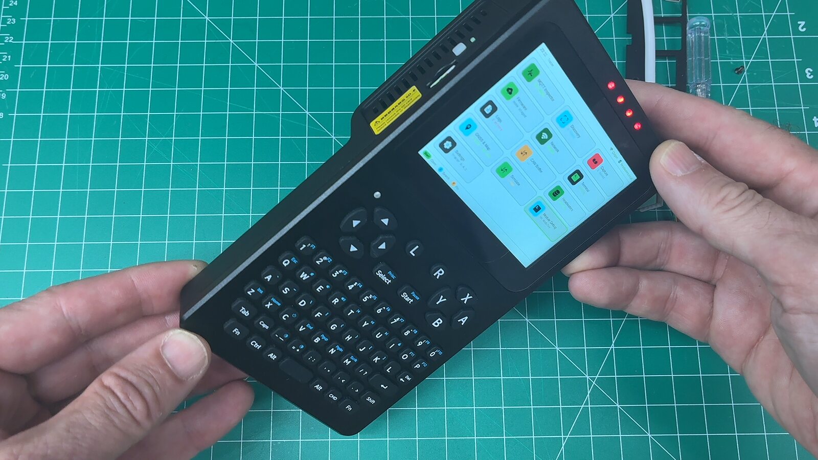 A Waveshare PocketTerm35 handheld held in two hands, showing the Tracer launcher with a grid of coloured diagnostic tiles on its 3.5 inch screen, above a full QWERTY keyboard and a D-pad
