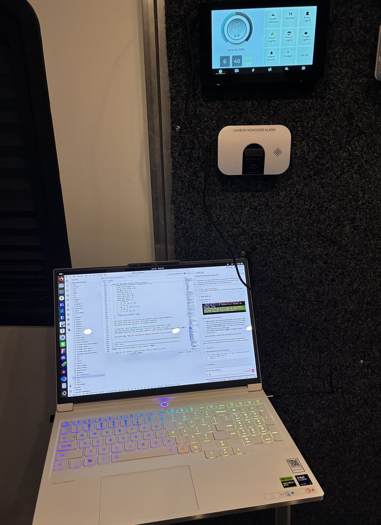 A laptop open on the floor of a travel trailer, screen full of firmware code, below a wall mounted TrailCurrent touchscreen showing lighting and climate controls and a carbon monoxide alarm